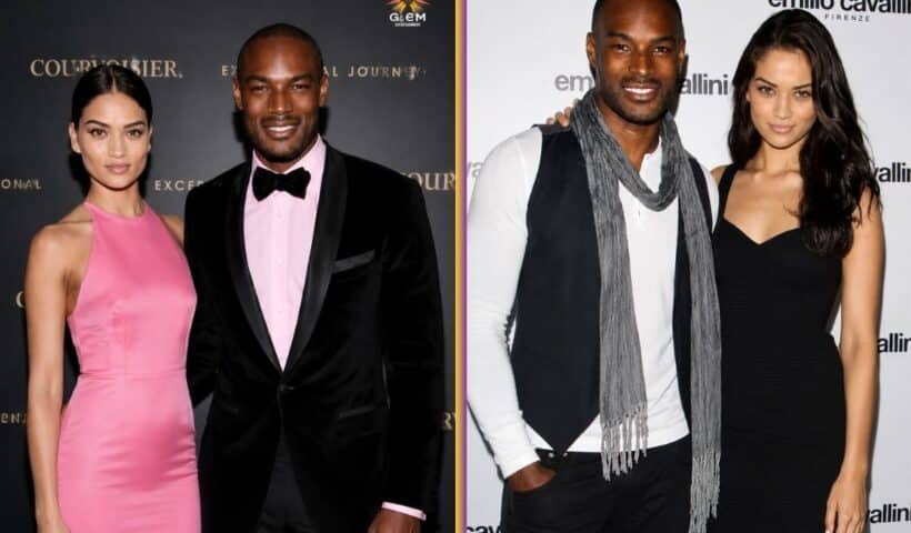 A well-dressed couple posing at a formal event in front of a branded backdrop. The man is wearing a white shirt with a dark vest and scarf, while the woman stands beside him in a fitted black dress. Both are smiling and facing the camera under bright lighting.