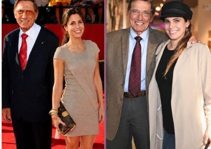 An older man wearing a brown checked blazer, light blue shirt, and maroon tie stands smiling beside a young woman in a black cap, black top, and beige trench coat. They pose together in a warmly lit art gallery, with framed artwork displayed along the walls in the background. The man has his arm around the woman, and both are looking directly at the camera.